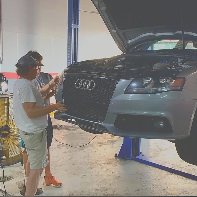 Two people working at the front of a car raised on a lift in the bay.